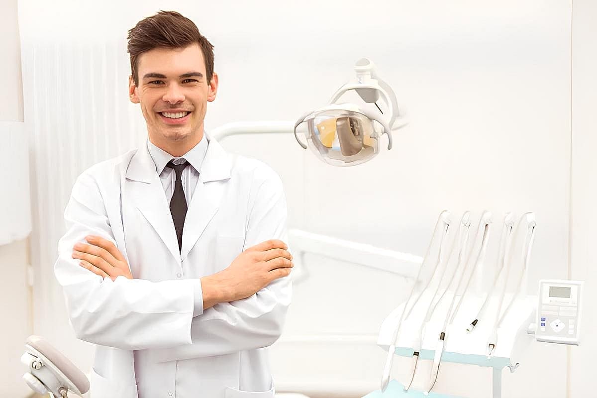 In Kingwood TX, a dentist in a white coat stands confidently with arms crossed in a dental office. Visible dental equipment showcases expertise in procedures like jaw surgery.